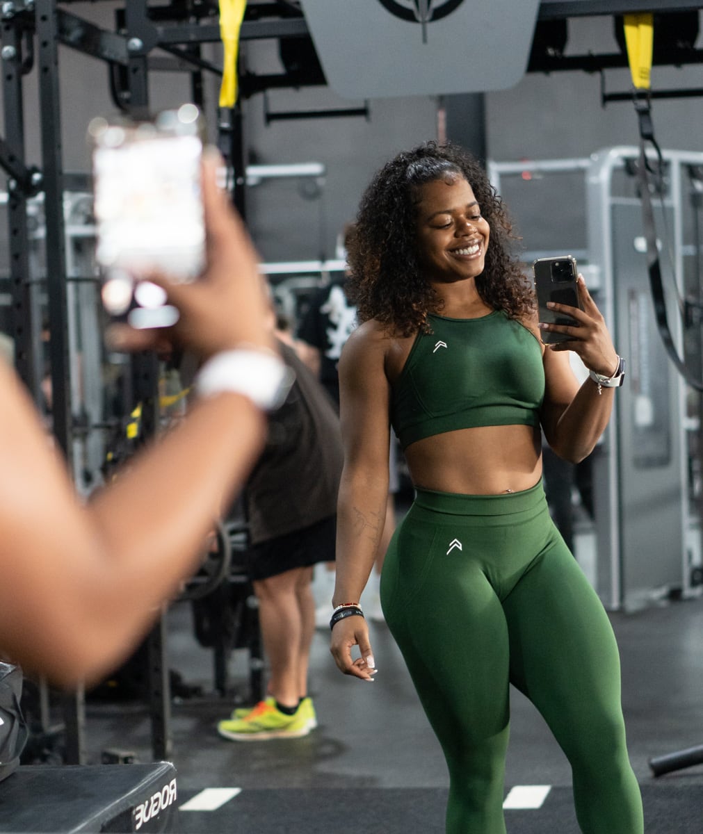 Coach on a green workout outfit is taking a selfi facing a mirror in the gym.